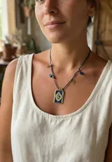 Collar con cordón trenzado azul oscuro con hilos dorados, dije rectangular dorado con un sagrado corazón en relieve sobre fondo azul esmaltado. Incluye cuentas de lapislázuli y dijes colgantes dorados con forma de estrella.
