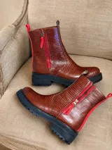 Botas de caña media con textura efecto crocco en color chocolate. Presentan cierre lateral con tirador en color rojo vibrante, ribetes rojos en la parte posterior y suela track negra de goma.