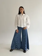 Blusa de brodery color blanco, con cuello redondo, mangas largas abullonadas con puños elásticos y cierre frontal con botones forrados. Presenta detalles de puntillas verticales en el frente.