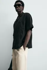 Camisa de corte relajado en color negro, confeccionada en tejido de algodón. Presenta cuello tipo bowling, manga corta y cierre frontal con botones. Cuenta con bordados lineales de hojas en la parte delantera.