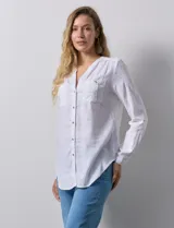 Camisa blanca de lino y rayón, con cuello a la base y escote en V abotonado. Tiene dos bolsillos con botón en el pecho y mangas largas regulables con presilla y botón.