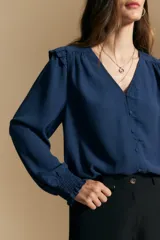 Camisa negra de manga larga con escote en V y cierre frontal de botones. Presenta detalles de volados en los hombros, frunces en la espalda y puños elastizados que le otorgan una silueta relajada y fluida.