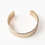 Brazalete rígido abierto de metal dorado con textura irregular y acabado martillado.