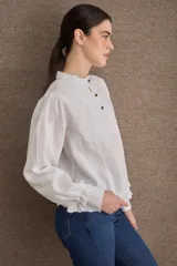 Blusa de lino color blanco con cuello mao, cierre frontal con botones, mangas largas abullonadas con puños ajustados y detalle de volado en el ruedo.