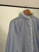 Camisa de rayas verticales celestes y blancas, con cuello alto y cierre frontal de botones. Presenta detalles de puntilla blanca en el cuello, a lo largo de la botonera y en los puños. Tiene mangas largas con terminación acampanada y diseño holgado.