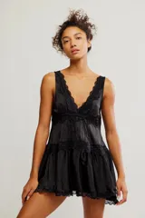 Mini vestido tipo slip de tela satinada negra, con escote en V profundo, diseño en capas con volados y detalles de encaje en los bordes. Corte estilo babydoll, sin mangas y silueta amplia.