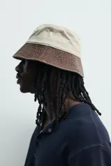Sombrero bucket confeccionado en tejido de algodón con efecto lavado, diseño bicolor en tonos beige claro y marrón oscuro. Presenta ala ancha con pespuntes y un bordado frontal en contraste.