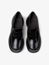 Mocasín de piel vacuna color negro, con suela de plataforma y tacón grueso. Presenta un diseño clásico con antifaz en el empeine y logo de la marca en el talón.