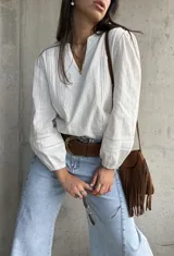 Blusa blanca de mangas largas abullonadas con puños elásticos y detalles de encaje y bordados sutiles. Presenta escote en V con tiras decorativas y diseño holgado.