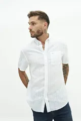Camisa de manga corta confeccionada en mezcla de lino y algodón, con diseño de rayas verticales sutiles en tono celeste sobre fondo blanco. Presenta cuello camisero con botones, cierre frontal abotonado y un bolsillo en el pecho con el logo de la marca bordado.