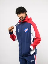 Campera deportiva Umbro del Club Nacional de Football, con cierre frontal, capucha ajustable con cordones y diseño en bloques de color azul, celeste y blanco. Presenta el logo de la marca y el escudo del club bordados en el pecho.