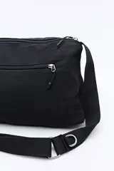 Cartera tipo bandolera de nylon color negro, con correa ajustable y detalle de hebilla metálica lateral. Presenta un diseño minimalista con cierre superior y logo sutil en la parte inferior.