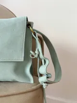 Bandolera de cuero color beige claro, con solapa frontal, cierre metálico de broche y correa regulable con detalles ondulados.