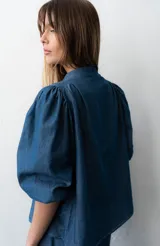 Blusa de denim liviano de corte asimétrico en tono azul, con mangas abullonadas, cuello redondo y cierre lateral con lazos para atar y botones.