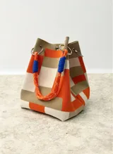 Cartera tipo bucket con diseño de bloques de color en tonos naranja, beige y crema. Confeccionada en lona resistente, cuenta con una manija de cuerda náutica naranja con detalles en azul y herrajes metálicos plateados.