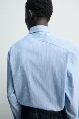 Camisa de algodón celeste con rayas verticales finas en blanco, de corte relajado (relaxed fit), cuello solapa y manga larga con puño abotonado.
