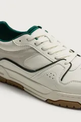 Zapatilla deportiva de estilo retro con diseño de paneles en combinación de materiales. Presenta una base de color blanco con detalles en verde oscuro y gris, y una suela de goma con volumen en tono caramelo.