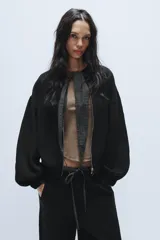 Chaqueta de punto negra con cuello redondo, cierre frontal con cremallera metálica y detalle de lazada en tejido a cuadros en contraste. Presenta mangas largas abullonadas con puños elásticos y bajo elástico.