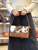Cartera de cuero marrón con solapa de pelo vacuno en tonos marrón y blanco, con cierre metálico giratorio y correa ajustable para el hombro.