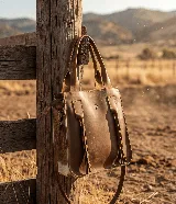 Cartera de cuero vacuno color marrón, hecha a mano en Uruguay. Presenta paneles laterales de cuero con pelo en tonos blanco y marrón. Tiene doble asa superior, una correa ajustable y desmontable, y detalles de flecos de cuero en el frente. Incluye forro interior y cierre.