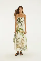 Vestido largo blanco de algodón con tirantes finos, escote recto y corte amplio en línea A. Presenta un estampado posicional tropical con palmeras, hojas de banano y arquitectura colonial.