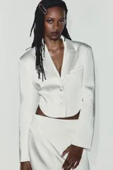 Blazer crop satinada de color blanco, con solapas, manga larga, cierre frontal con botones y detalle de abertura en la espalda.