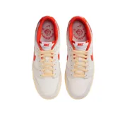 Championes Nike Dunk Low con diseño de paneles en tonos gris claro y blanco, destacando el swoosh lateral y detalles en el talón y cuello en color rojo vibrante. Presenta cordones en tono crema y una suela con efecto envejecido.