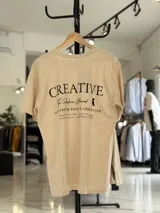 Remera oversize color beige, con diseño estampado en la espalda que incluye tipografía grande y texto descriptivo en negro.