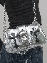 Cartera de hombro color plateado metalizado con diseño rectangular, cierre frontal con hebilla y apliques de cruces negras en los laterales. Incluye una cadena decorativa plateada y correa ajustable.