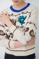 Buzo de tejido de punto en color beige con estampado de motivos florales y abstractos en tonos marrones. Presenta cuello redondo con borde en color azul vibrante, mangas largas y puños ajustados.
