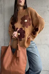 Sweater de punto grueso color camel, con diseño de flores grandes aplicadas en tonos marrones y centro crema. Presenta cuello redondo alto, mangas largas con hombros caídos y terminaciones acanaladas.