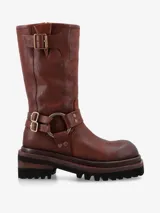 Bota de caña media de cuero marrón envejecido, estilo biker, con suela track gruesa y dentada. Presenta una correa ajustable con hebilla metálica dorada en la parte superior de la caña y otra correa con hebilla y argolla metálica grande en el tobillo.