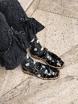 Sandalias tipo pescador de cuero negro con acabado brillante, diseño de tiras entrelazadas, cierre con hebilla metálica lateral y suela plana con borde en tono madera.