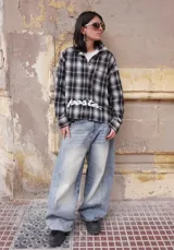 Camisa de manga larga con corte boxy fit, confeccionada en algodón a cuadros en tonos negro y gris. Presenta un bolsillo frontal en el pecho y el texto 'posta.' estampado en color blanco en la parte delantera.