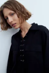 Vestido corto negro con cuello camisero, mangas largas, cierre frontal con cordones y bolsillos de parche en el pecho y las caderas.