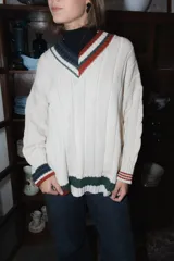 Buzo tejido de punto color terracota con diseño de rayas verticales en relieve. Presenta cuello en V con detalles de rayas en azul marino, blanco y negro, y puños acanalados a juego. Incluye un pequeño bordado en el pecho.