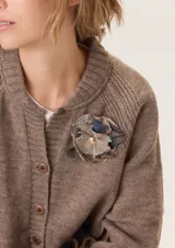 Prendedor artesanal con forma de flor, confeccionado en una mezcla de tejidos con texturas variadas en tonos marrón, beige y azul. Presenta un detalle central decorativo en color dorado.