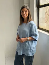 Blusa celeste de corte holgado, con cuello redondo, mangas tres cuartos con trabilla y detalles de alforzas en el frente.