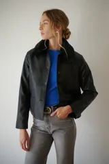 Chaqueta corta color beige con cuello de piel sintética negra, cierre frontal de botones y bolsillos laterales.