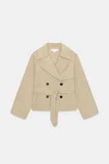 Trench corto de color beige, con cuello de solapa, manga larga con hombreras y trabillas con botones. Cuenta con cierre frontal cruzado con botones oscuros y cinturón ajustable del mismo tejido.