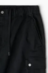 Pantalón cargo negro de corte holgado, confeccionado en tejido de algodón. Presenta cintura con elástico, bolsillos laterales al bies, bolsillos de fuelle con botones a presión en las piernas y pliegues en las rodillas.