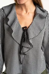 Blusa de manga larga con estampado de cuadros vichy en blanco y negro. Presenta un cuello con volados amplios y un detalle de lazo negro en el centro del escote.