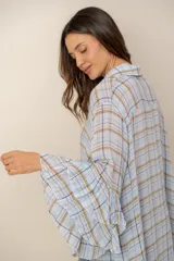Camisa de manga larga con diseño a cuadros en tonos celestes, marrones y blancos. Presenta corte asimétrico con largo irregular, más larga en la parte trasera, y mangas amplias con volados escalonados.
