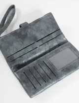 Billetera plegable de cuero vegano con acabado metalizado en tono azul grisáceo. Cuenta con una correa de mano (strap) integrada, cierre principal y un broche metálico decorativo en el frente. Incluye logo de la marca en la parte inferior.