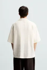 Camisa de calce relajado confeccionada en tejido de algodón color beige. Presenta un diseño de jacquard con líneas verticales desflecadas, cuello tipo bowling, manga corta y cierre frontal con botones.