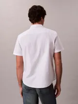 Camisa blanca de manga corta, confeccionada en tejido Oxford de algodón orgánico con elastano. Presenta cierre frontal con botones, cuello abotonado y logo de Calvin Klein Jeans bordado en el pecho.