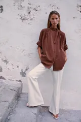 Camisa bordó de cuello alto ajustable con doble hebilla y cierre frontal con botones ocultos por solapa. Tiene mangas cortas con trabillas y diseño holgado.