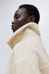 Campera de sherpa suave con cuello elevado, cierre frontal y bolsillos laterales de parche. Presenta un diseño de hombros caídos y corte holgado.