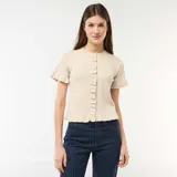 Camiseta de punto color beige con cuello redondo, cierre frontal de botones y mangas cortas con volantes. Presenta detalles de volantes en la botonera y en el ruedo.