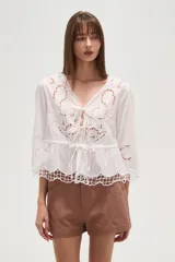 Blusa blanca de algodón con bordados tipo broderie anglaise, escote en V con lazos ajustables en el centro del busto y peplum con terminación festoneada. Mangas tres cuartos.